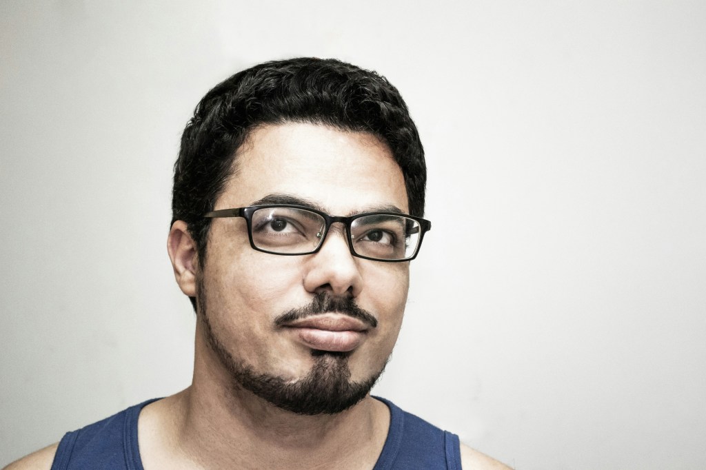A close-up portrait of a man with glasses and a goatee, wearing a blue tank top, against a plain white background.