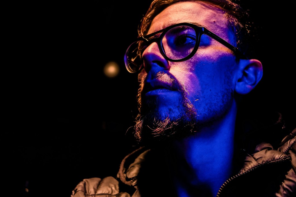 Close-up portrait of a young man with glasses, styled beard, and illuminated by dramatic blue lighting against a dark background.