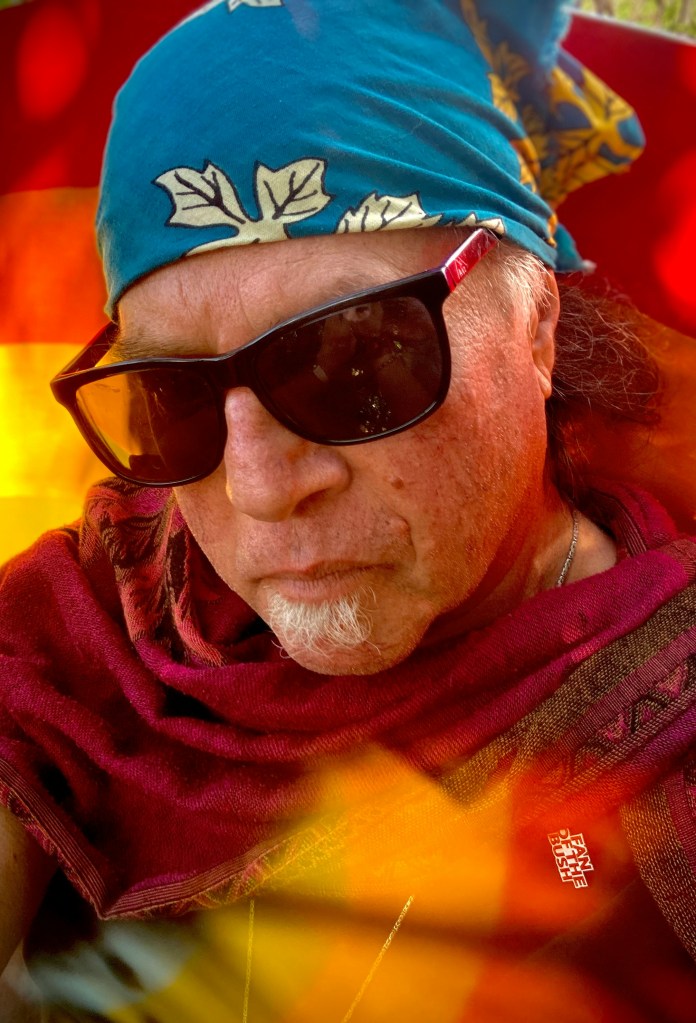 A close-up portrait of a person wearing sunglasses and a colorful bandana, with a warm, artistic background.