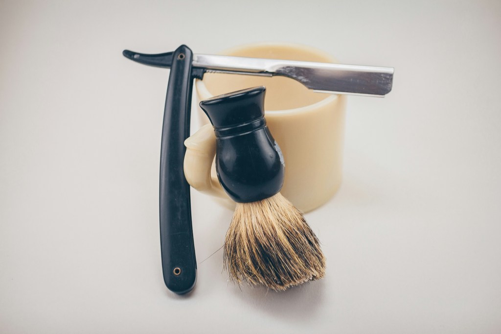 A shaving brush and a straight razor placed on a flat surface next to a ceramic holder.