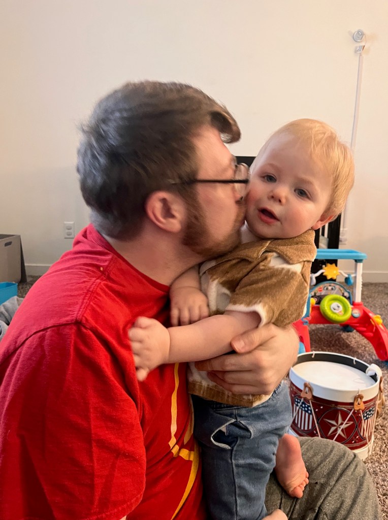 A young child in a brown shirt is being hugged by a person in a red shirt, both smiling. The background contains toys and a carpeted floor.